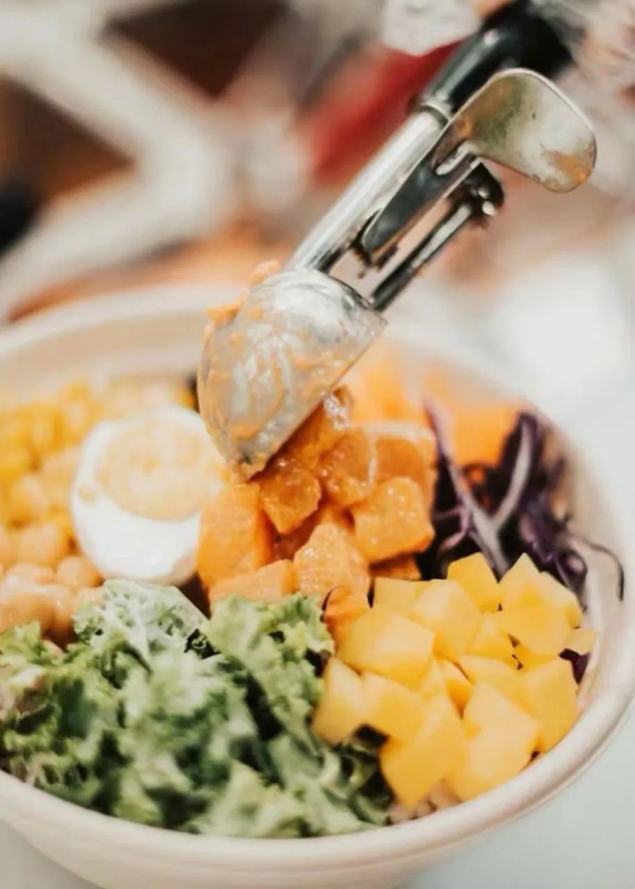 Metal scoop adding salmon to a poke bowl with corn, egg, mango, and greens at Natural Kitchen Wilton, a Fast Casual Restaurant in Wilton