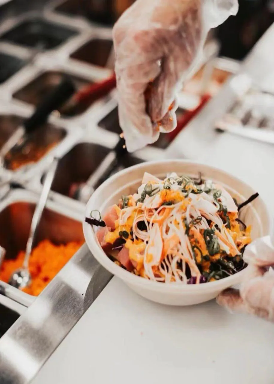 Hands adding crispy toppings to a poke bowl at Natural Kitchen Wilton, a Fast Casual Restaurant in Wilton
